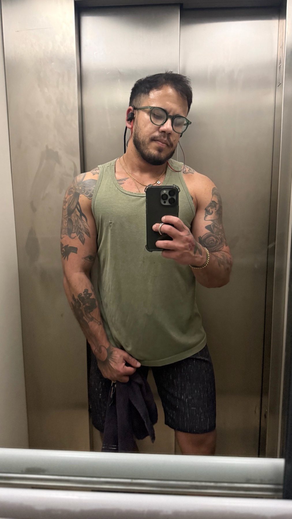 A man takes a mirror selfie in an elevator, showing off his muscular physique and tattoos. He is wearing a tank top and shorts.