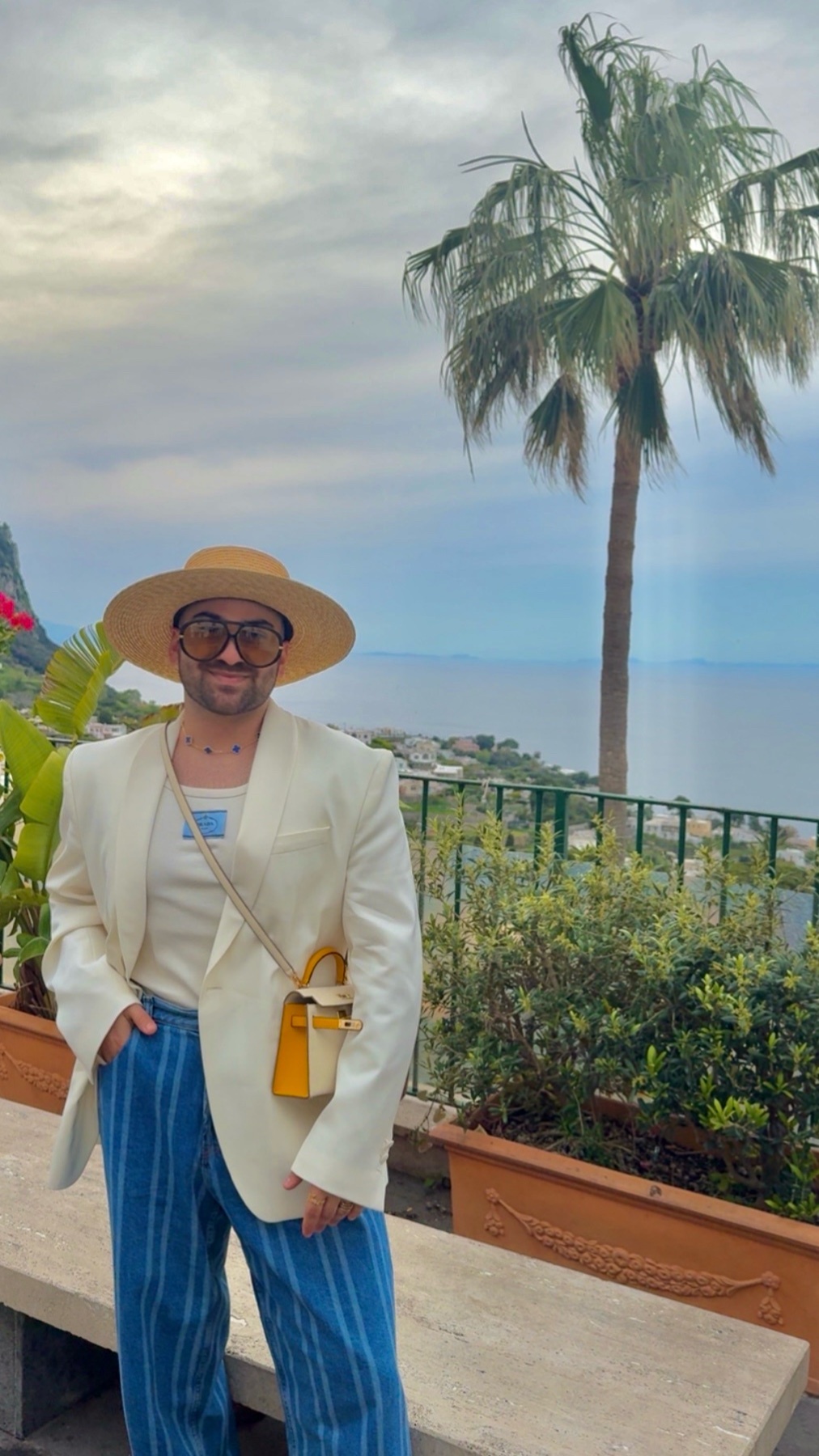 A man poses for a photo in a stylish outfit, including a wide-brimmed straw hat, a cream-colored blazer, and blue and white striped pants. He stands against a backdrop of lush greenery and the ocean, with a palm tree swaying gently in the breeze.