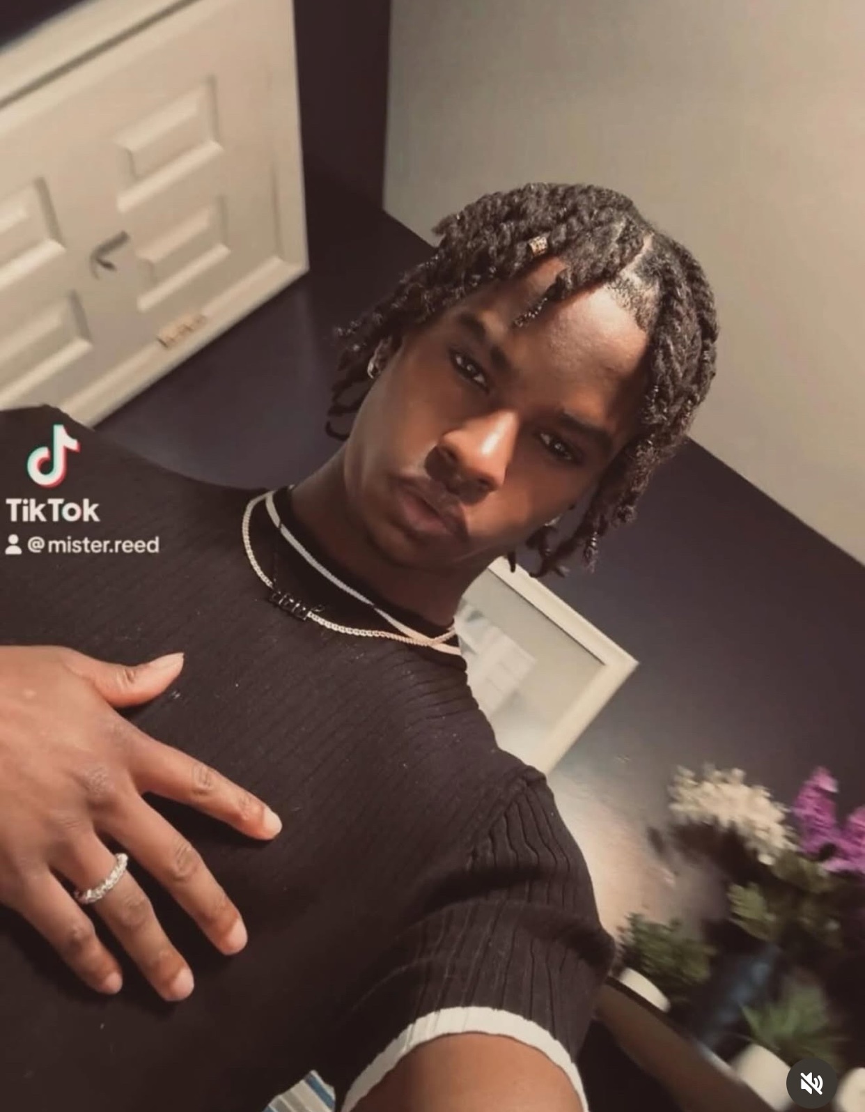 A man with dark braided hair poses for a selfie, wearing a black shirt and jewelry.