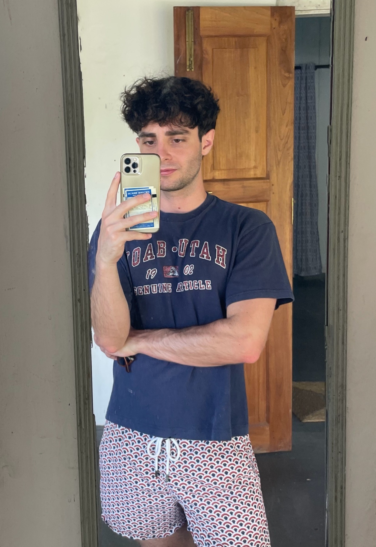 A young man with curly dark hair takes a mirror selfie in a room with wooden doors. He is wearing a navy blue t-shirt and patterned shorts.