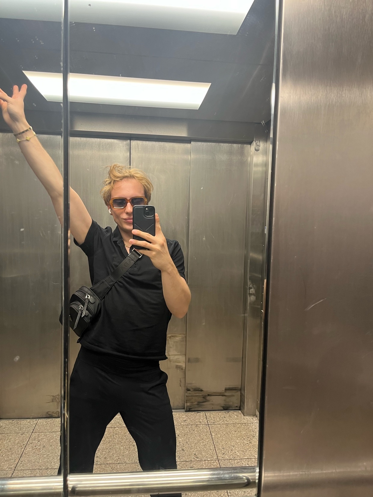A man takes a mirror selfie in an elevator, raising his arm. He is wearing a black outfit and sunglasses.