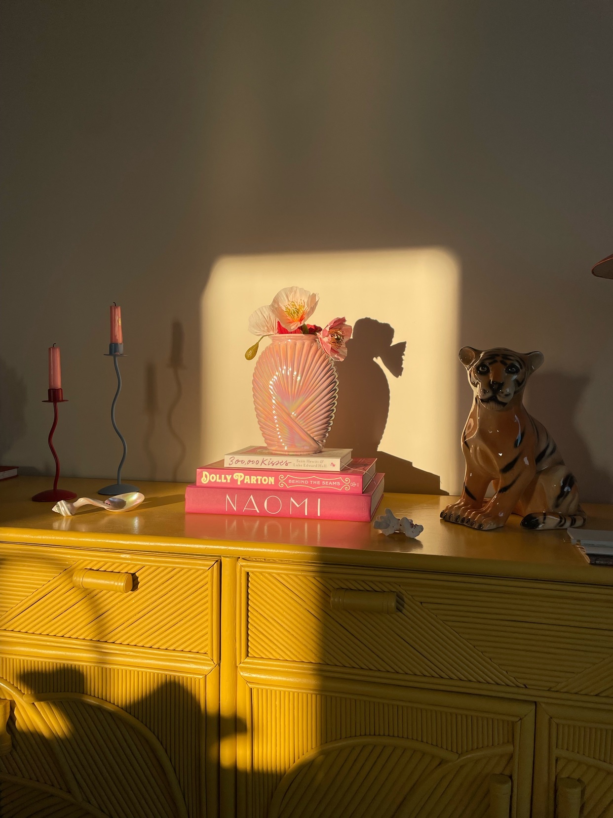 A vintage yellow cabinet is adorned with a pink vase, books, and a tiger figurine, bathed in warm sunlight and shadows.