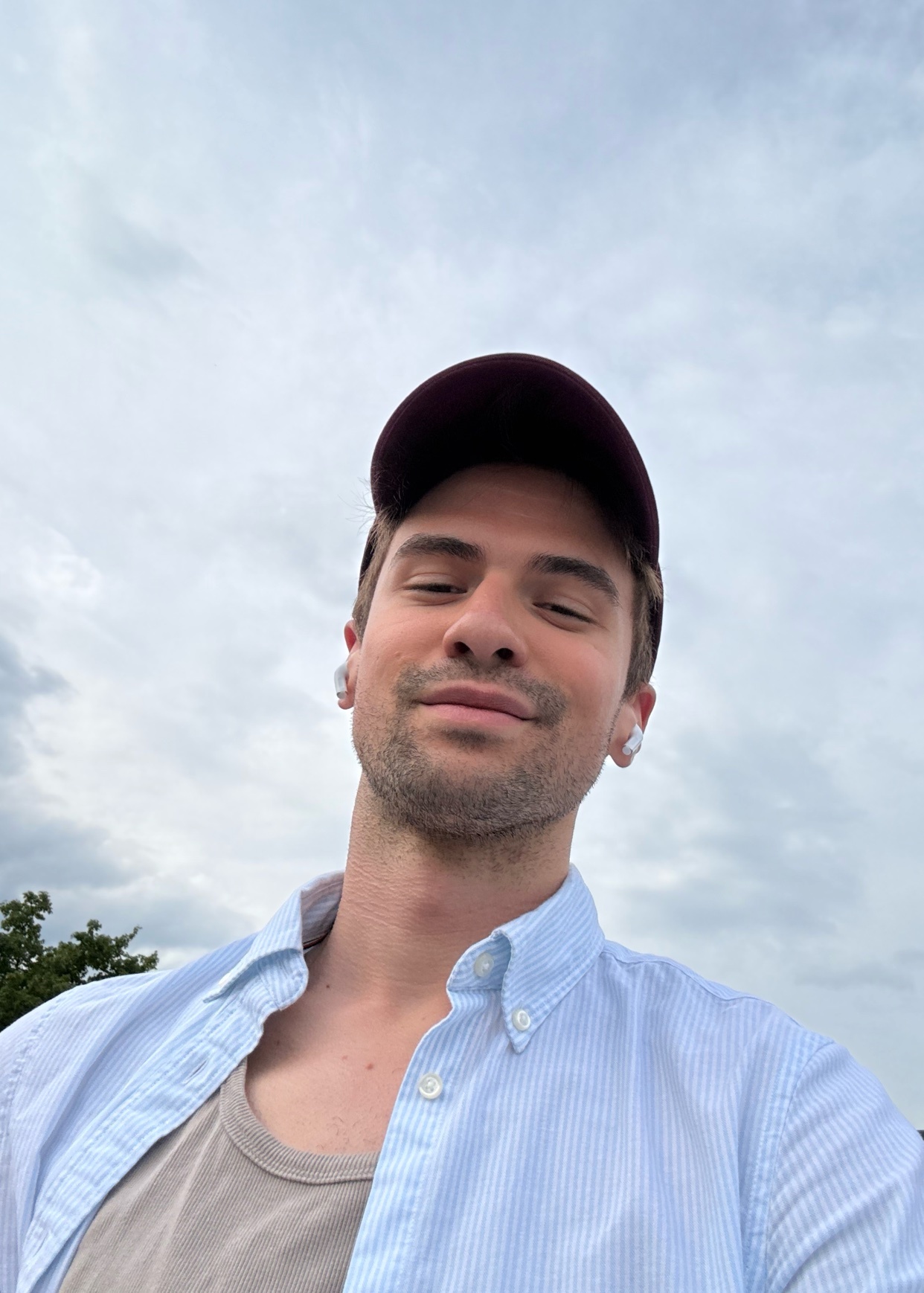 A man takes a selfie outdoors, wearing a cap and a blue shirt. The sky is visible in the background.
