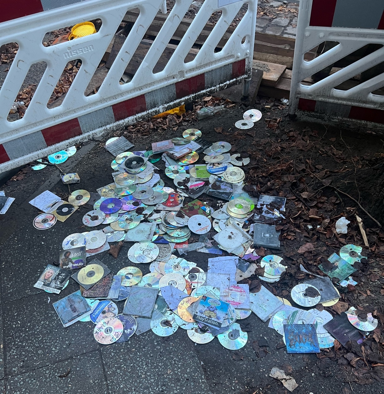 A pile of discarded CDs and their cases are strewn across a city street. The image captures a scene of urban waste and environmental concern.
