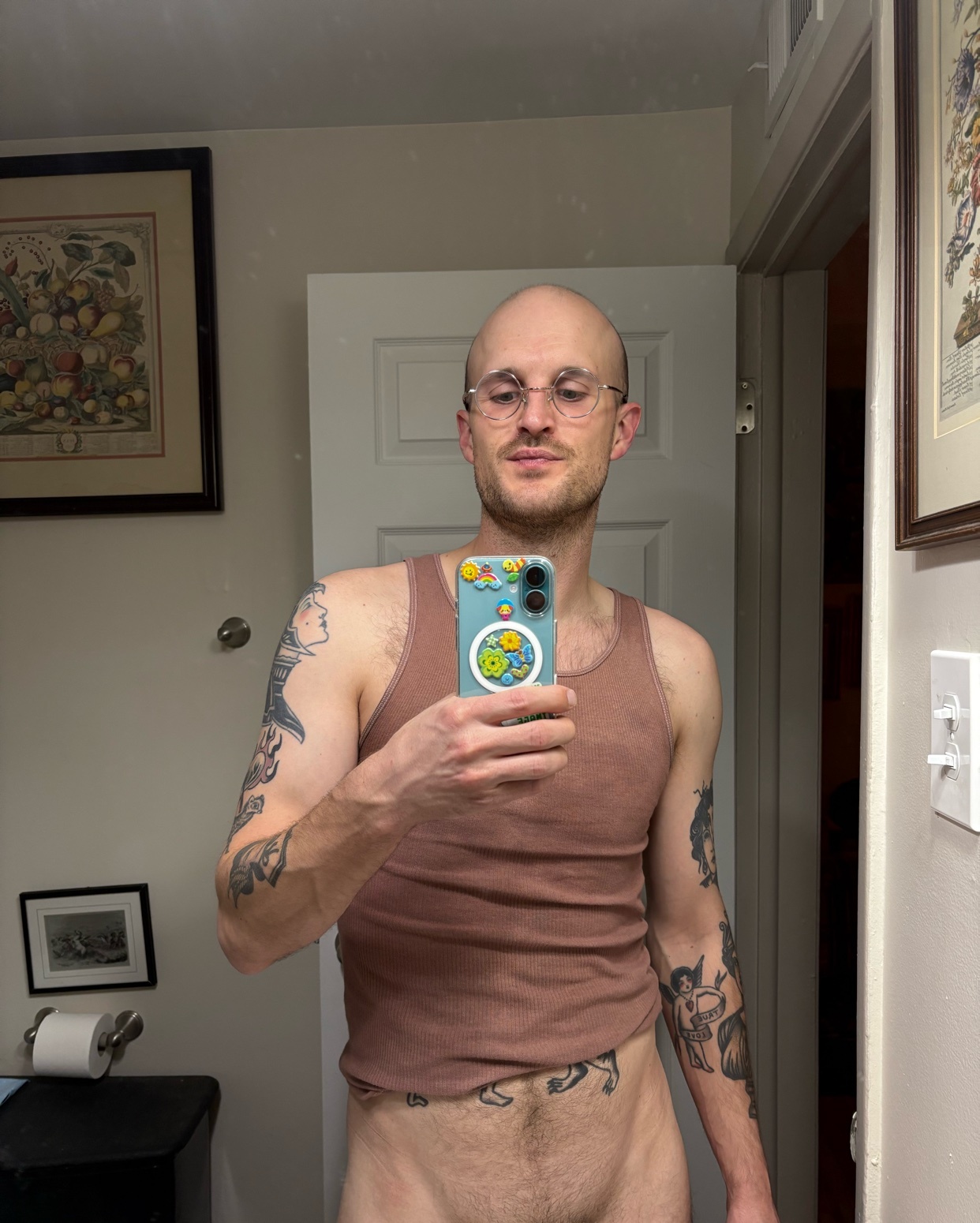 A man takes a mirror selfie, shirtless, in a bathroom. He has tattoos on his arms and is wearing glasses.