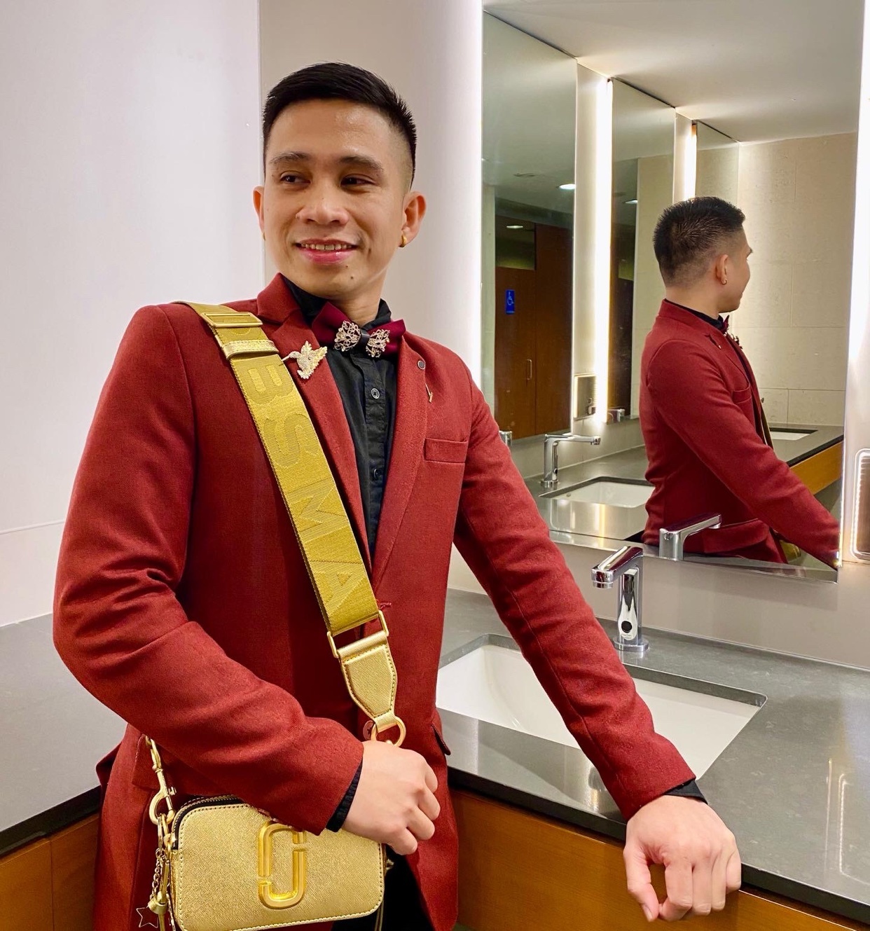 A man poses in a red suit with a gold shoulder bag. He is smiling and looking to the side.