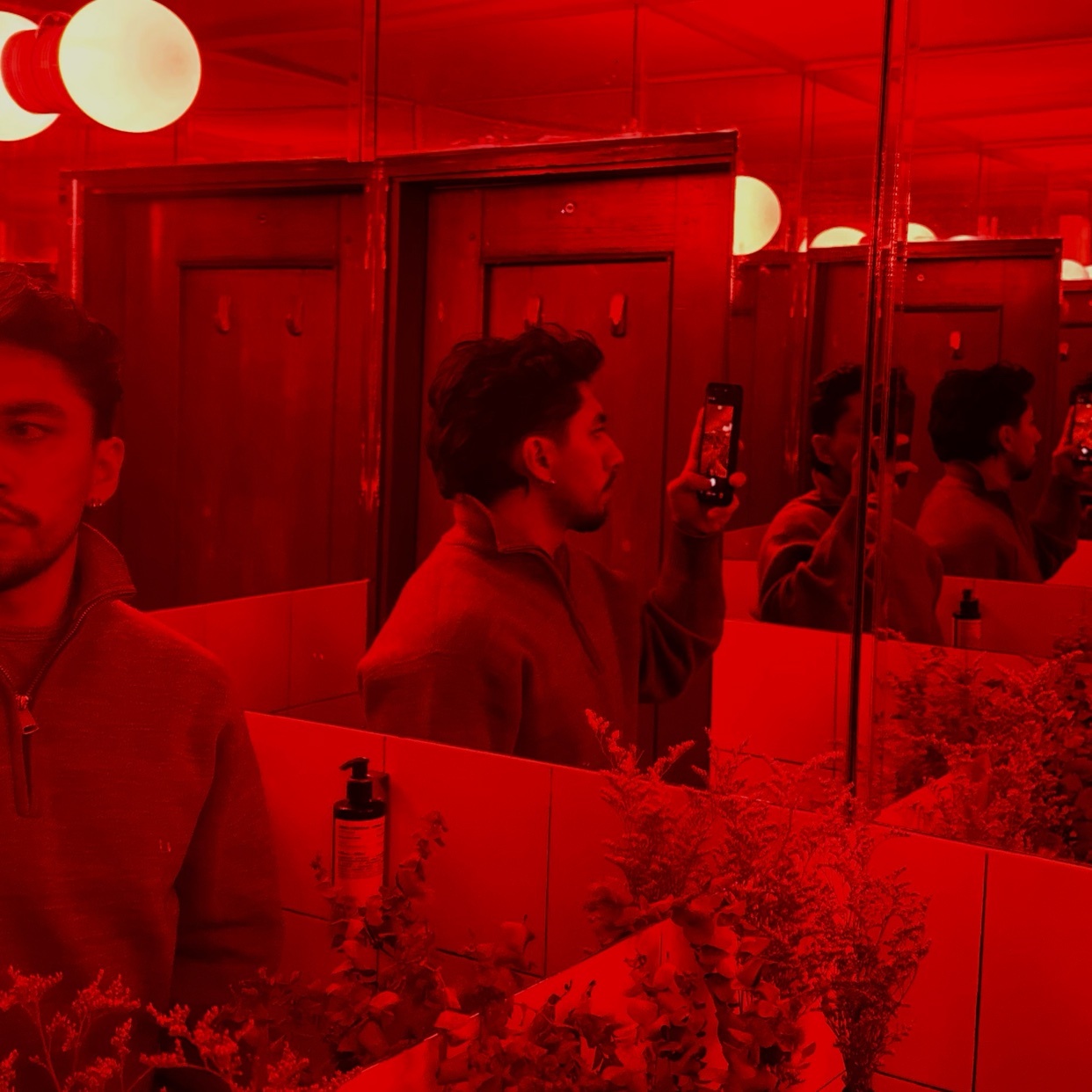 A man takes a selfie in a bathroom with red lighting. His reflection is visible in the mirror, surrounded by plants.
