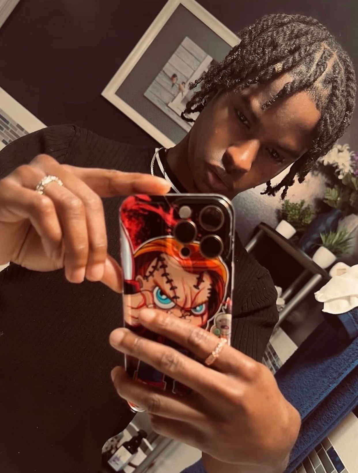 A young Black man with braided hair takes a selfie in a mirror. He is wearing a black sweater and several rings on his fingers. His phone case features a graphic of the horror character Chucky.