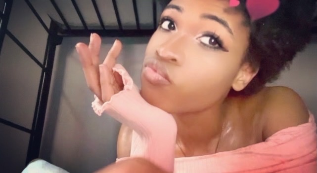 A young Black woman poses for a selfie, puckering her lips and resting her chin on her hand. She wears a pink top and has a heart filter over her head.