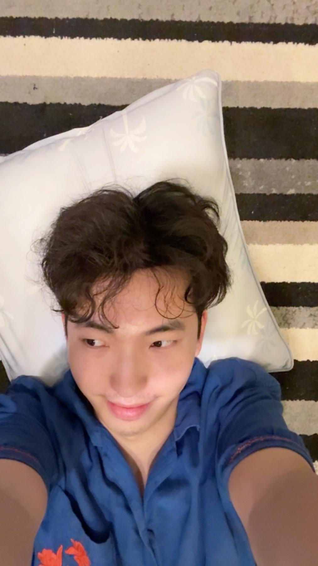 A man with curly dark hair lies on a pillow, looking towards the camera with a slight smile. He is wearing a blue collared shirt.