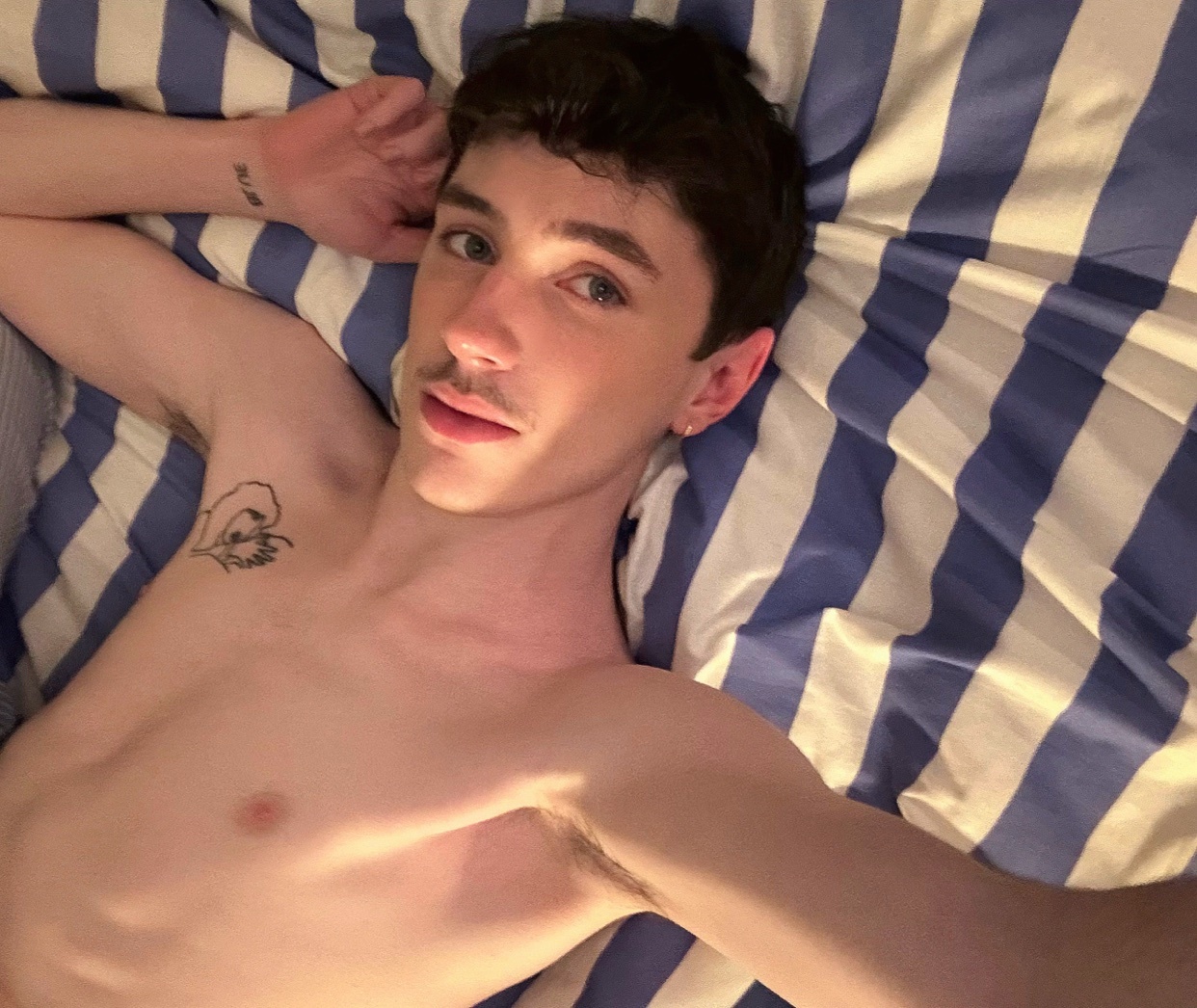 A shirtless man with dark hair and stubble lies on a bed with blue and white striped bedding. He has tattoos on his chest and arm.