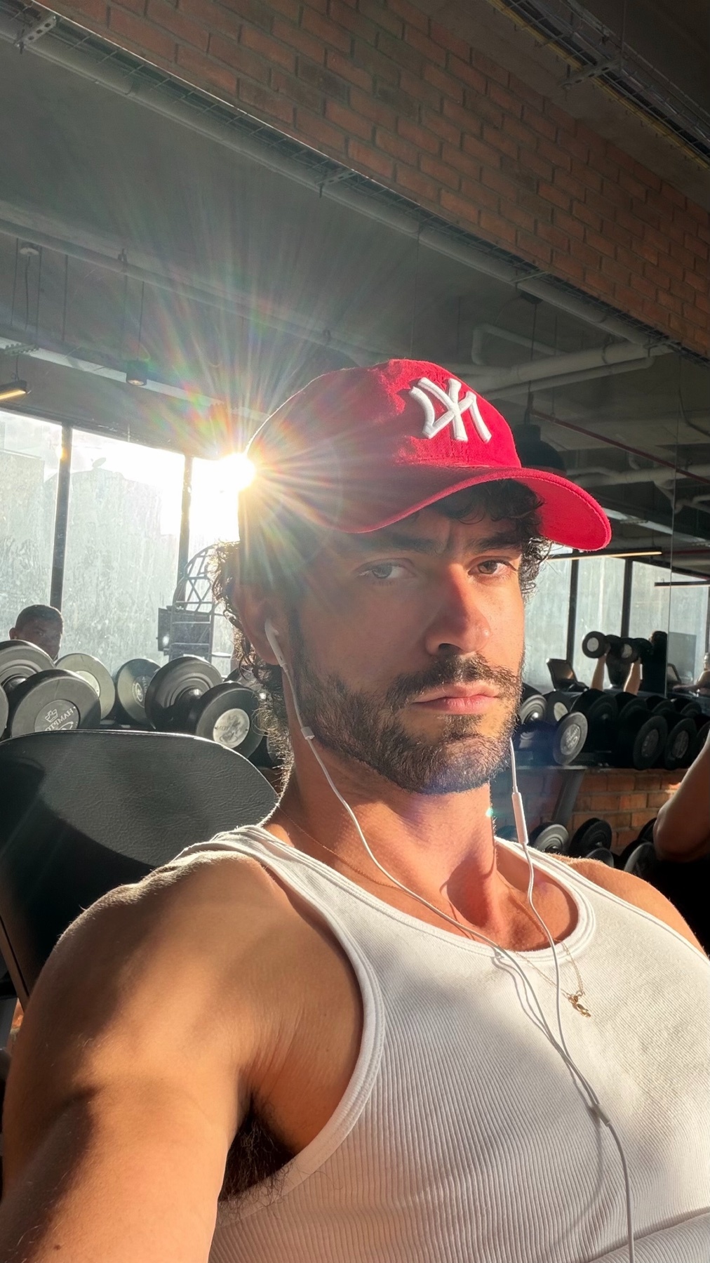 A man in a gym, wearing a red cap and a white tank top. He is shirtless and has a beard, with weights in the background.