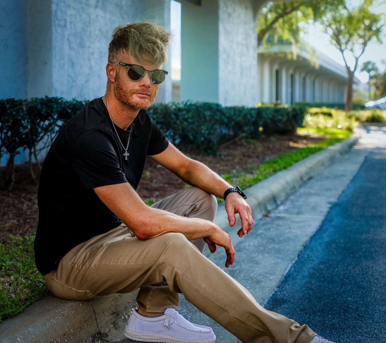 A man with blonde hair and sunglasses poses outdoors, sitting on a curb in a black t-shirt and khaki pants.
