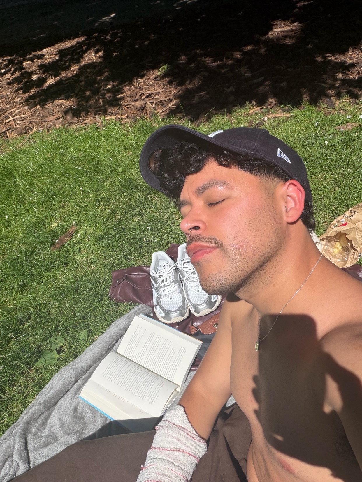 A shirtless man wearing a baseball cap and sunglasses relaxes in the sun in a park, with a book open beside him.