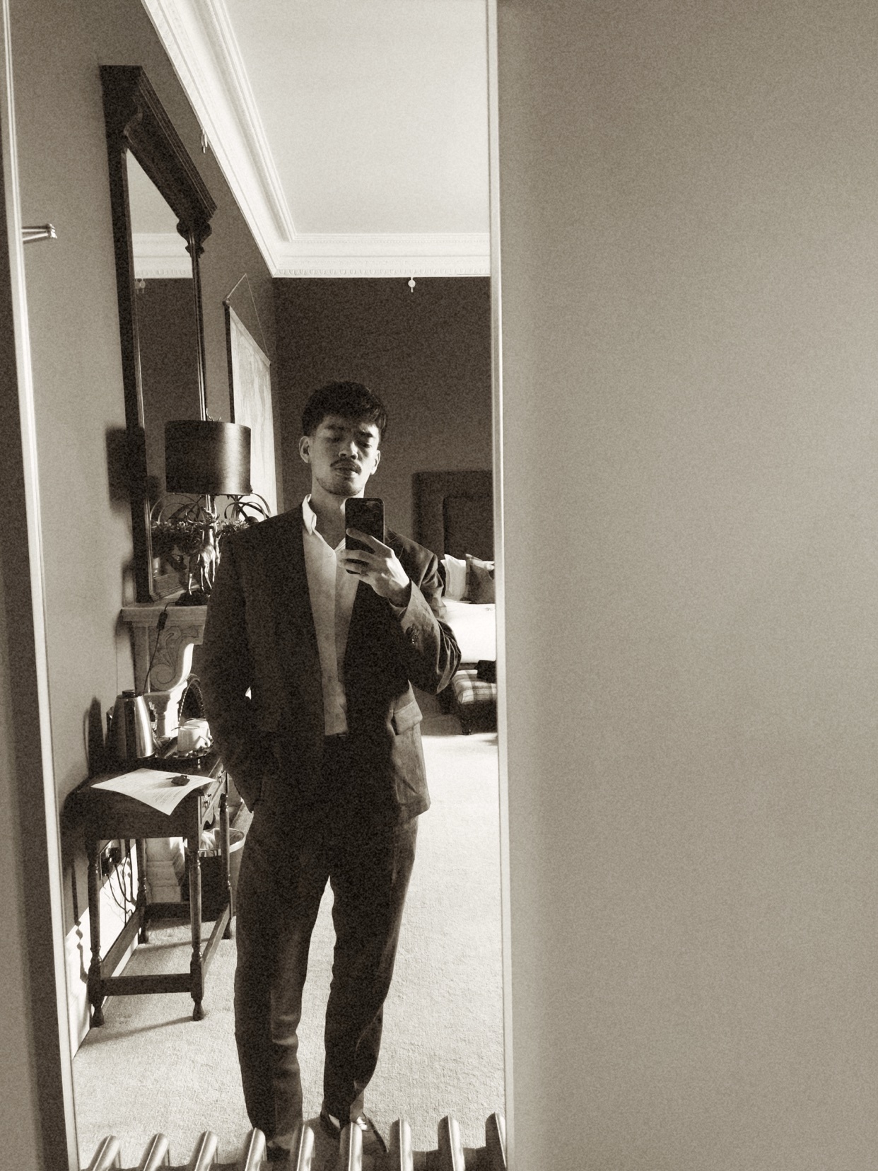 A man in a suit poses for a mirror selfie in a room, likely preparing for a wedding. He has stubble and a moustache, and his hand is in his pocket.
