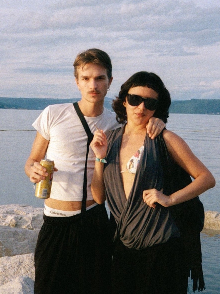A couple poses on a beach, the man holding a drink. The woman wears sunglasses and a draped top.