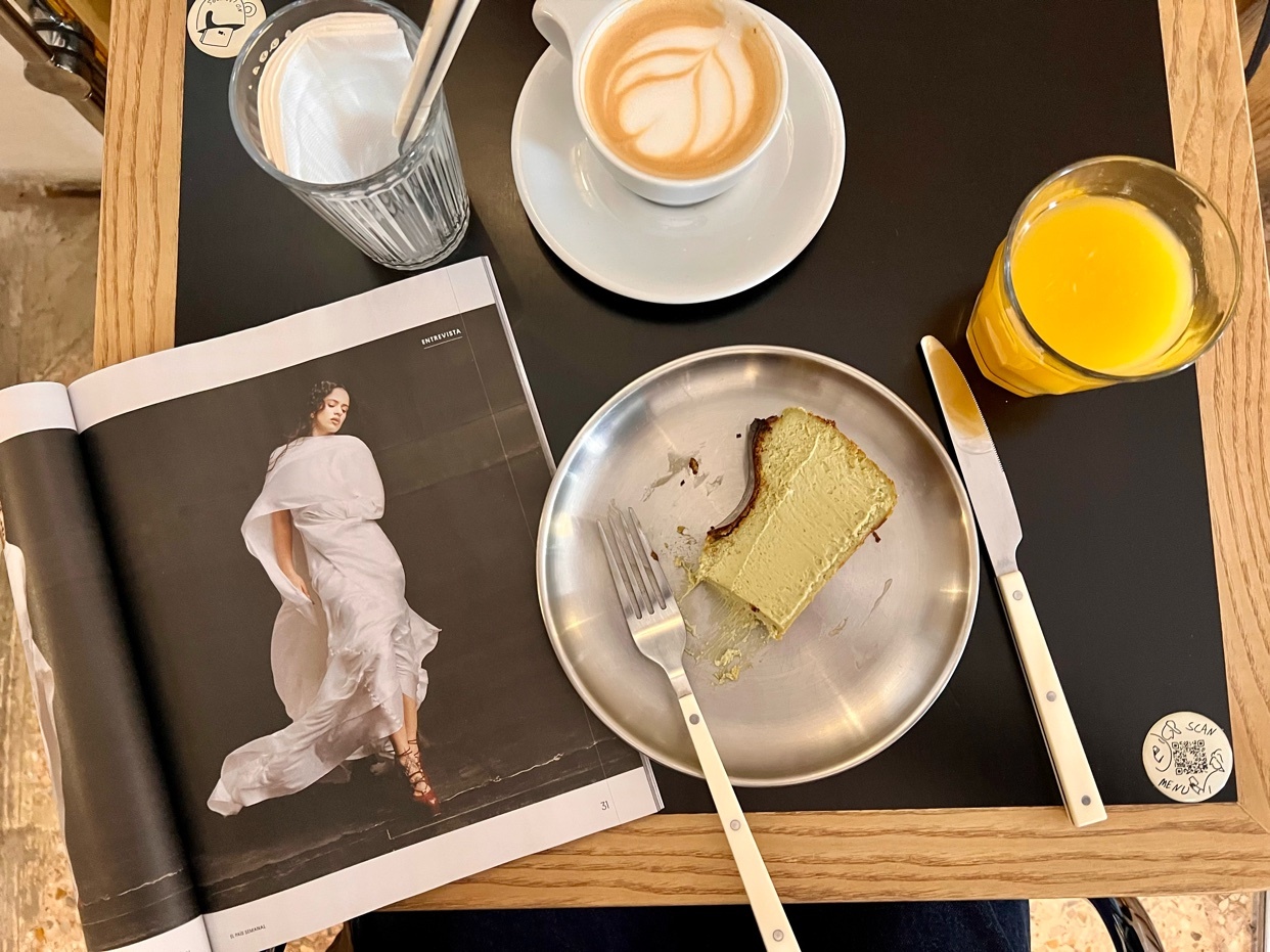 A flat lay of a table with coffee, cake, orange juice, and a magazine. The image has a clean and minimalist aesthetic.