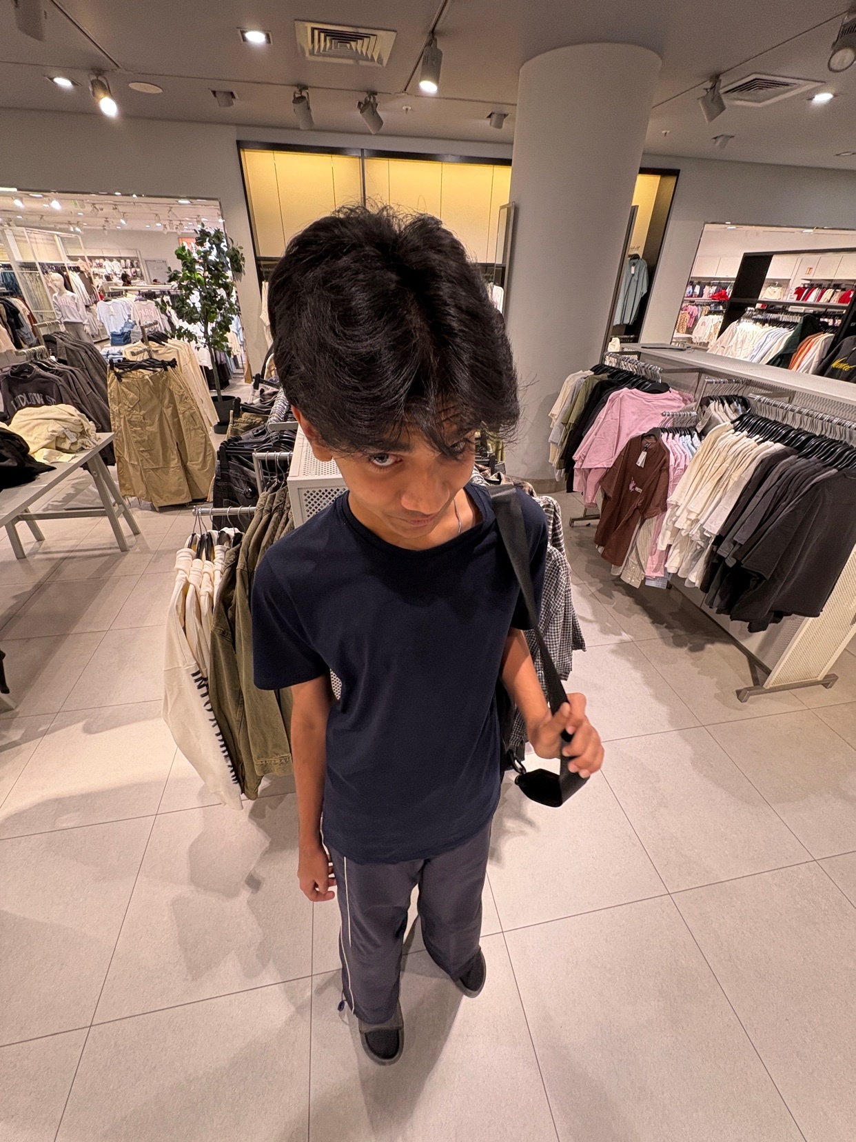 A young man poses in a clothing store, surrounded by racks of clothes. He wears a dark t-shirt and pants.