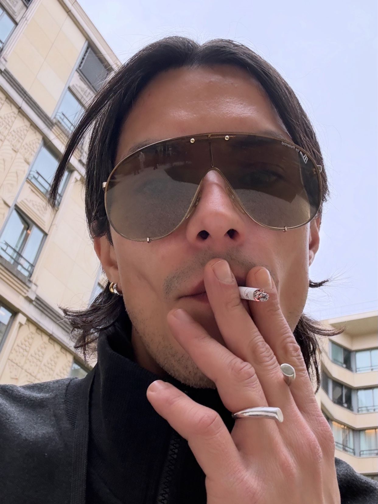A man is seen wearing large sunglasses and smoking a cigarette. The photo is a close-up portrait.