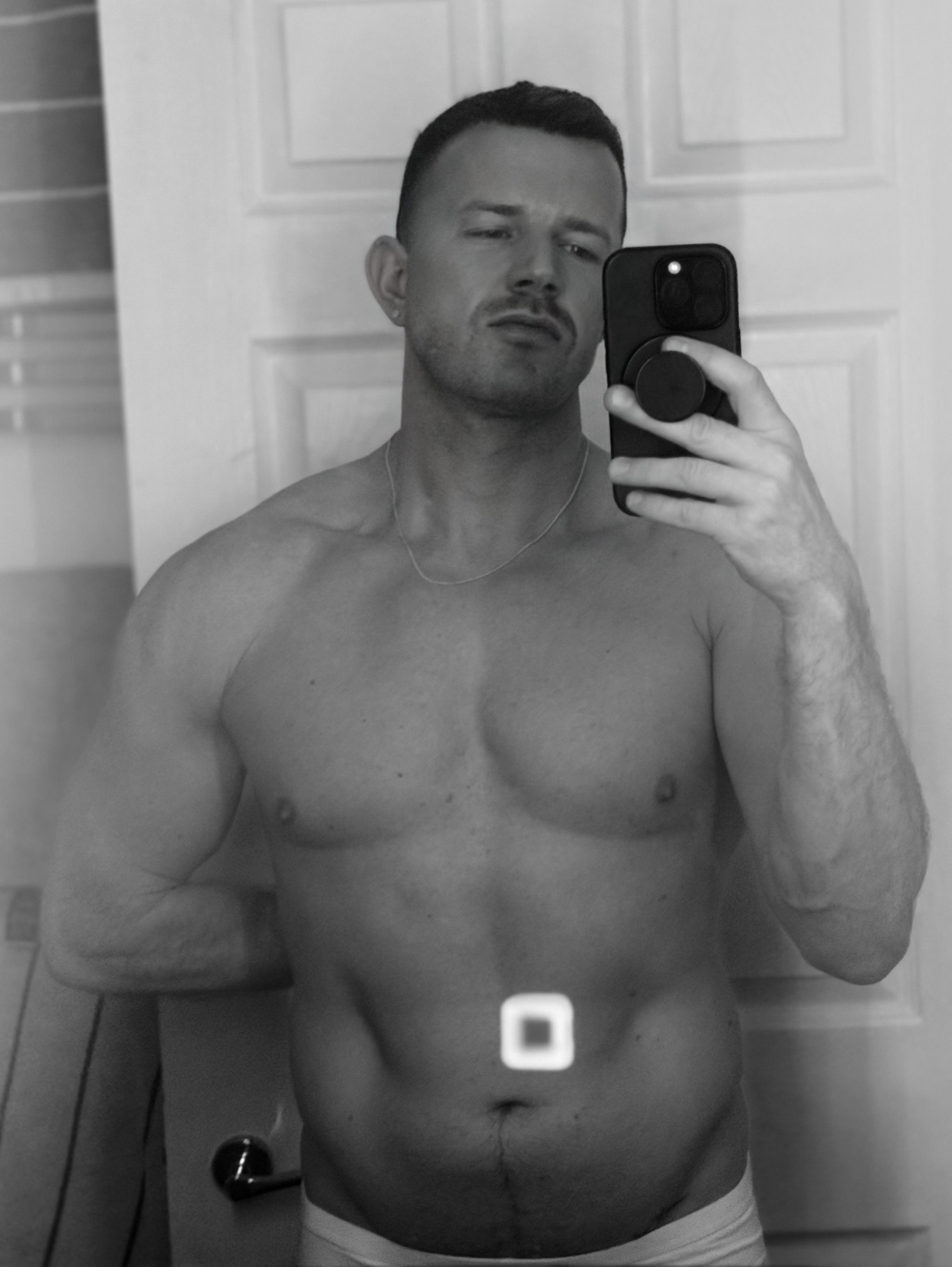 A shirtless man takes a mirror selfie, showcasing his muscular physique. The black and white photo emphasizes his physique and the intimate setting.