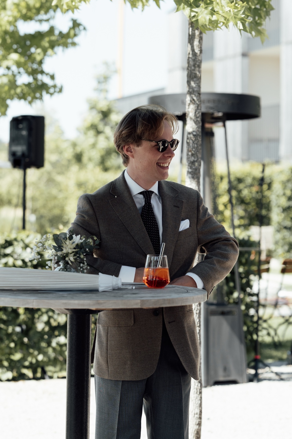 A man in a suit and sunglasses smiles while leaning on a table with a cocktail.