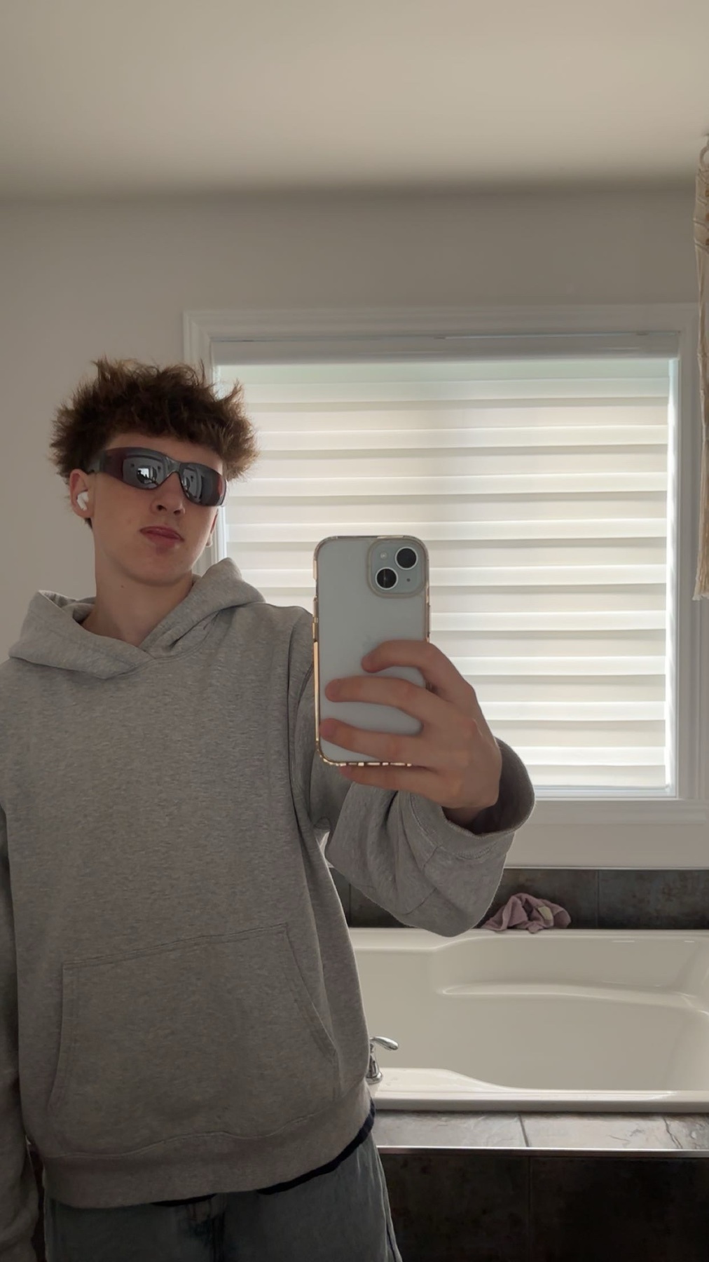 A young man takes a selfie in a bathroom, wearing sunglasses and a grey hoodie. He has curly hair and is wearing earbuds.