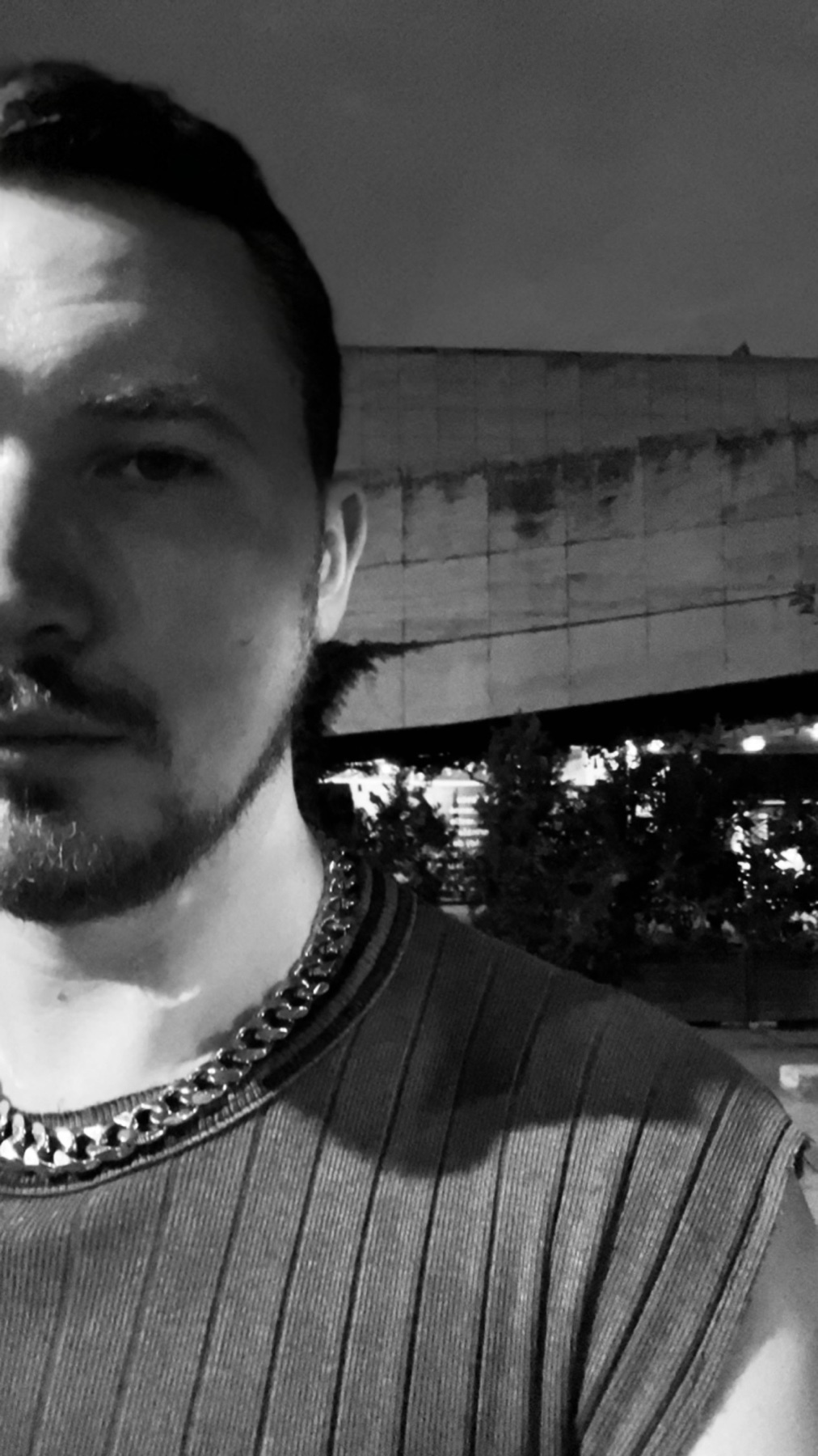 A black and white portrait of a man wearing a chain. The photo is taken at night with a cityscape in the background.