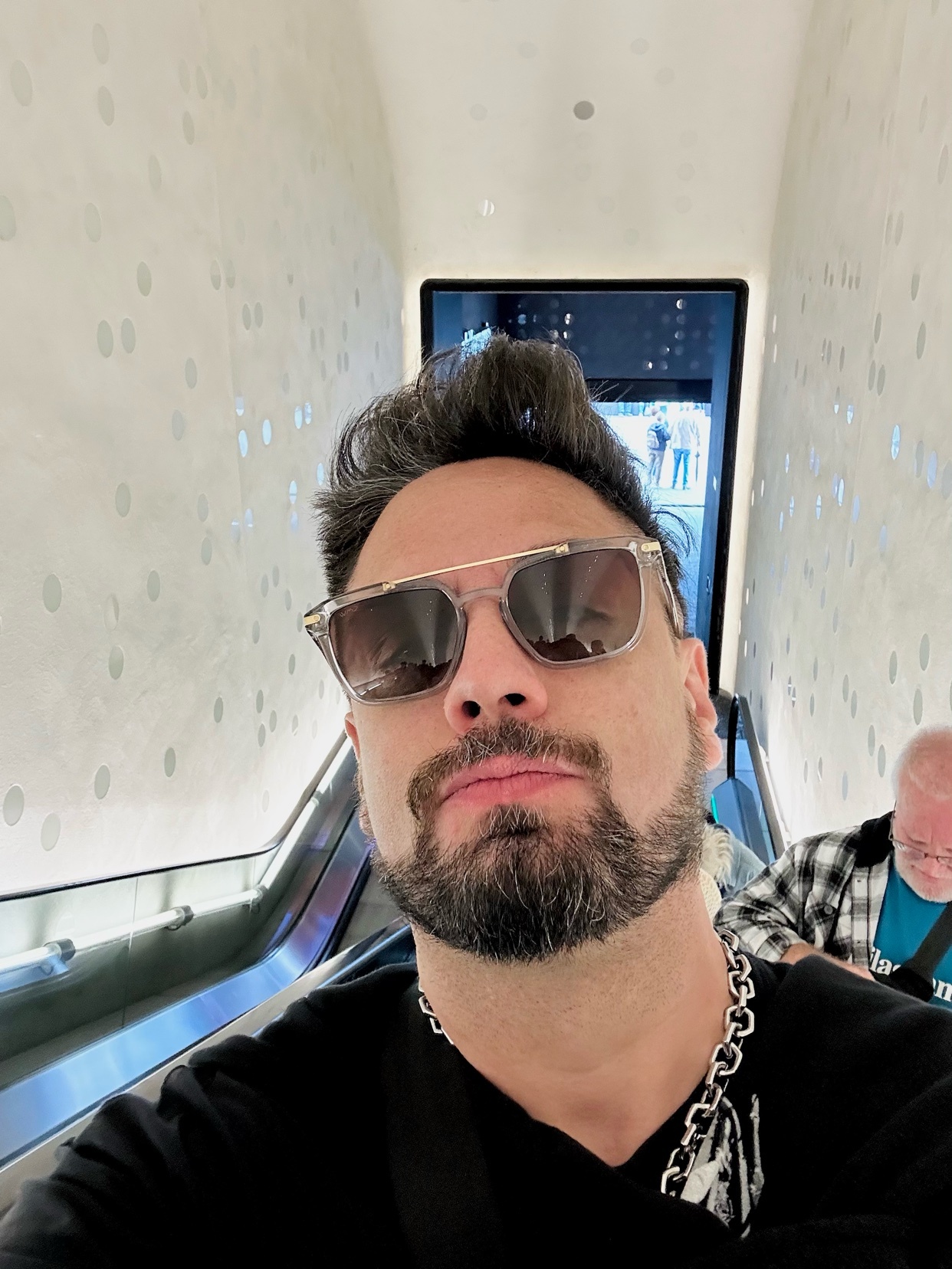 A man with a beard and goatee takes a selfie while wearing sunglasses and a chain necklace on an escalator.