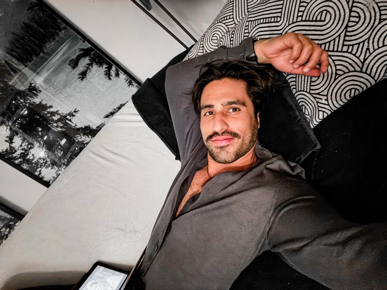 A man takes a selfie while lying down. He is shirtless and wearing a gray shirt.