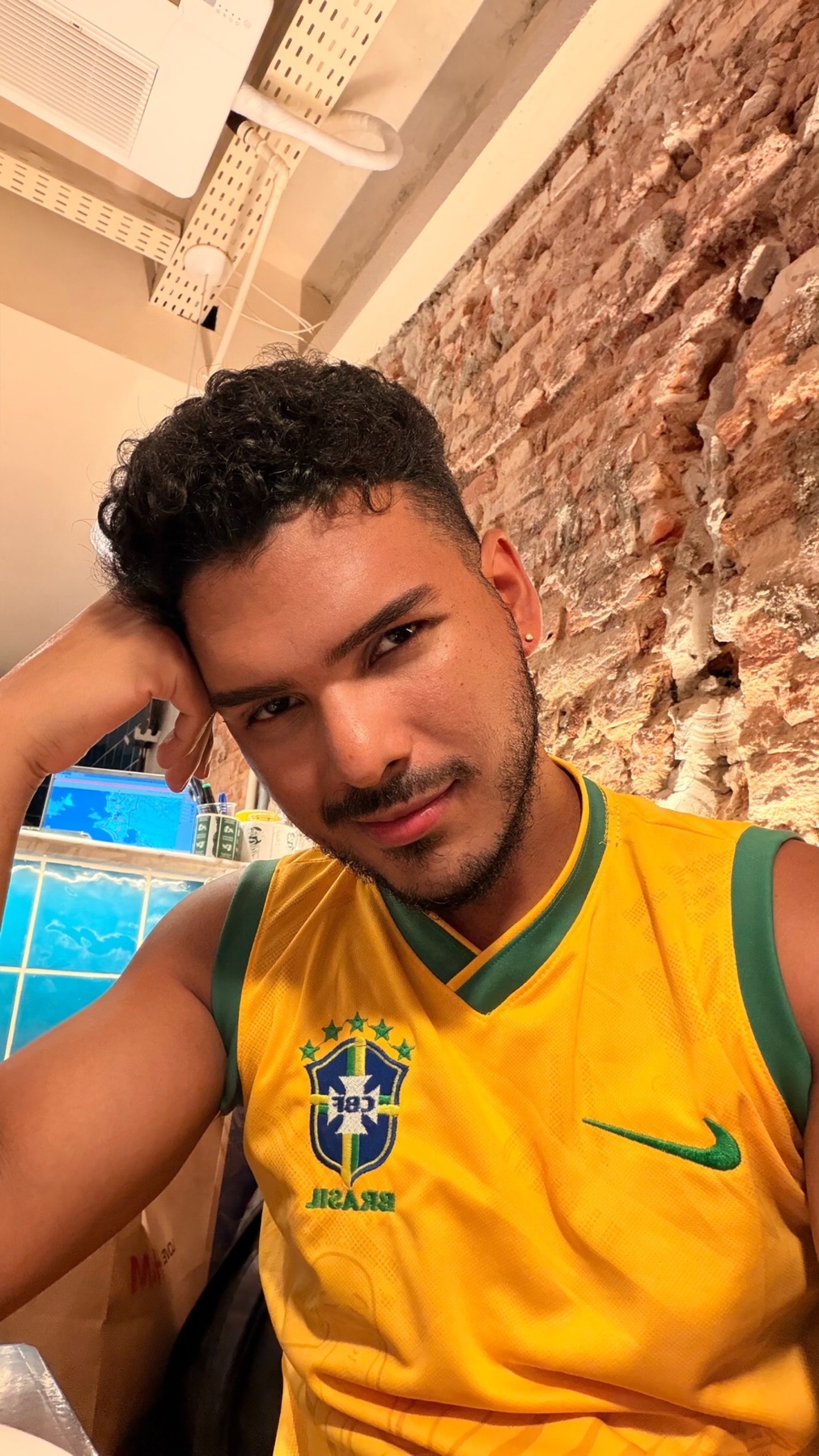 A man with curly dark hair and stubble poses for a selfie wearing a yellow Brazil national football team jersey.