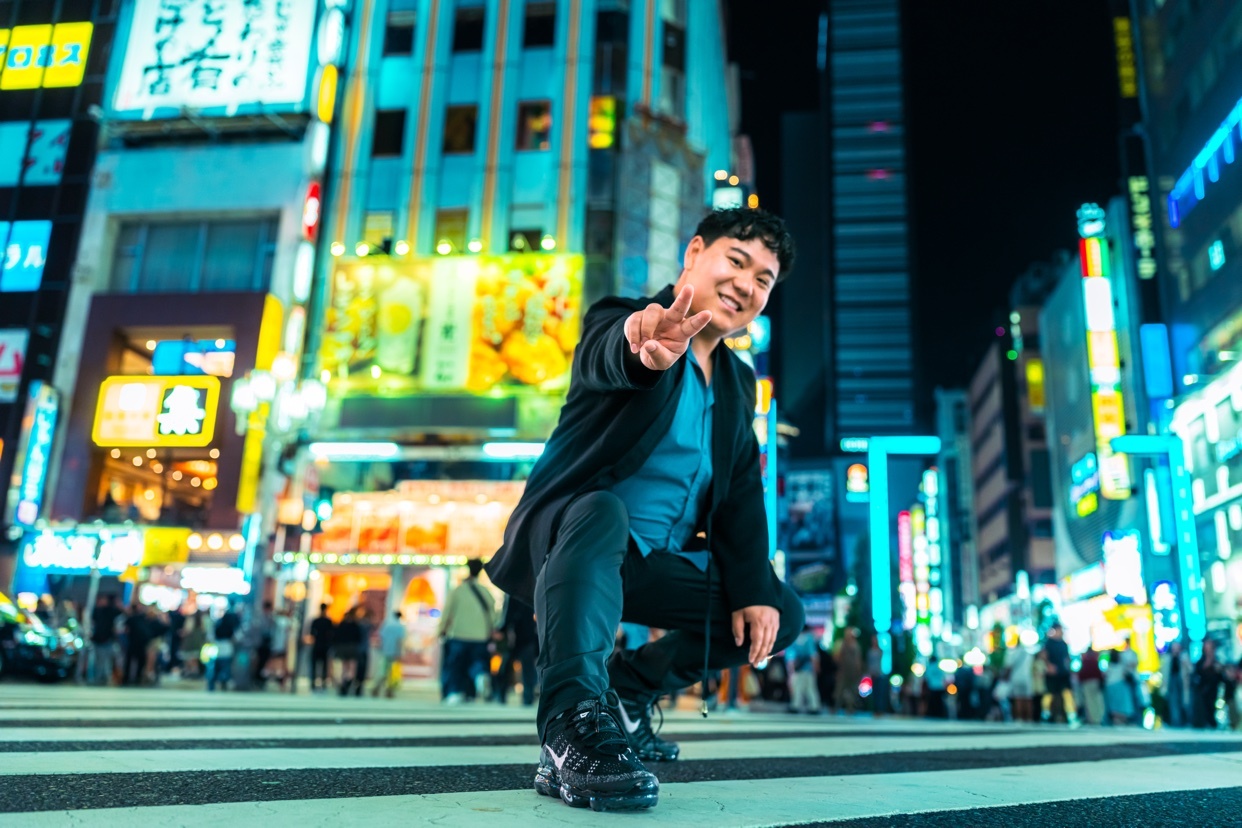 A man poses for a photo in a vibrant, neon-lit city street at night. He is crouching on a crosswalk, reaching his hand out towards the camera with a smile.