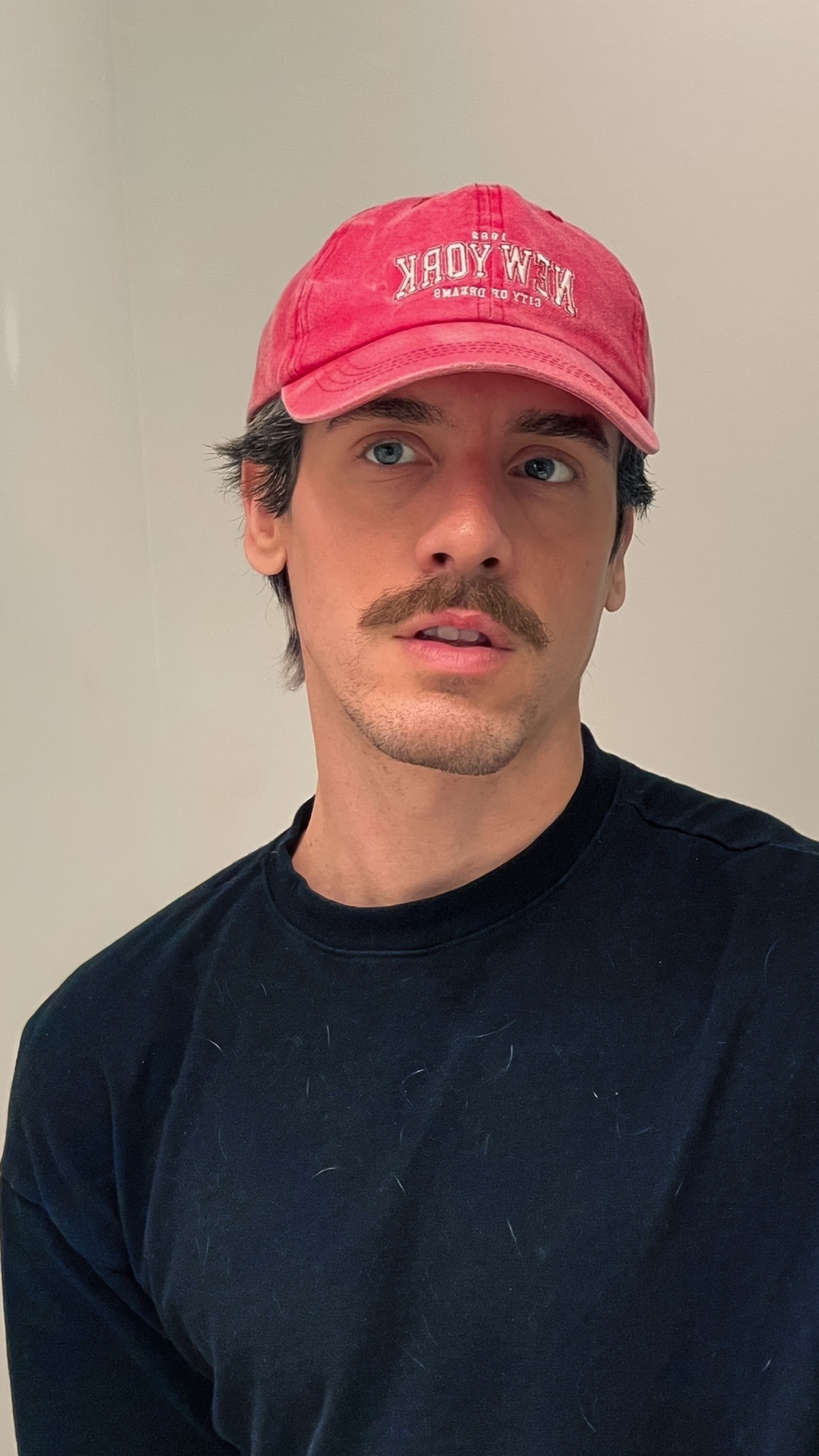 A man wearing a red New York cap and a black shirt. The man has a mustache and dark hair.
