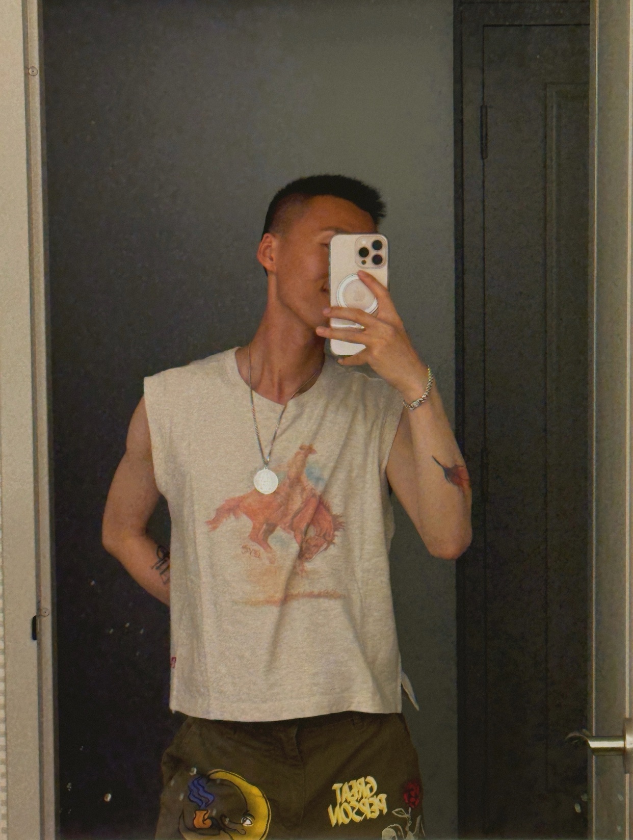 A man takes a mirror selfie, showcasing his outfit. He is shirtless, wearing a graphic t-shirt and cargo pants.