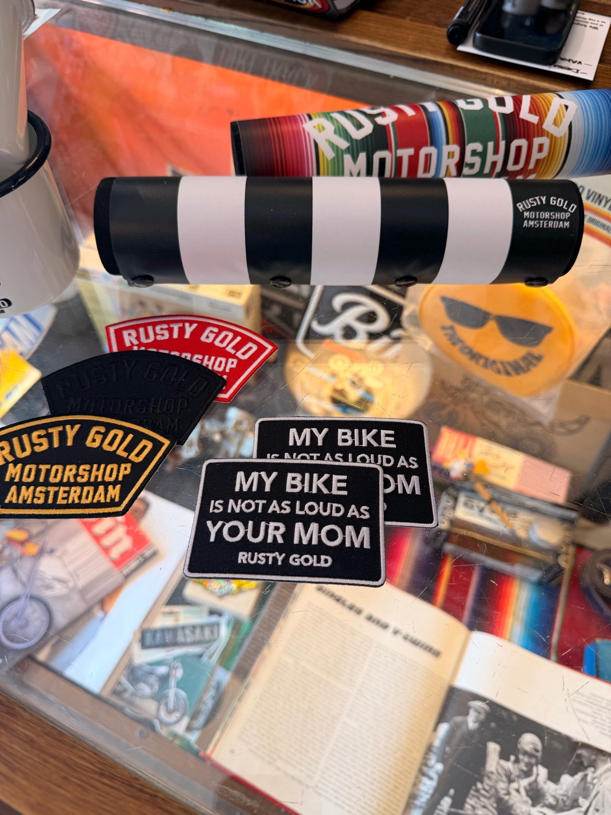 A collection of motorcycle-themed stickers and merchandise, including a black and white striped item and several stickers with the 'Rusty Gold Motorshop Amsterdam' logo. One sticker humorously states 'My Bike is Not As Loud As Your Mom'.