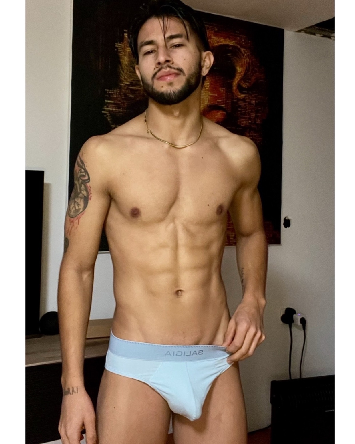 A man poses shirtless, wearing light blue underwear. He has a muscular physique and a tattoo on his arm.