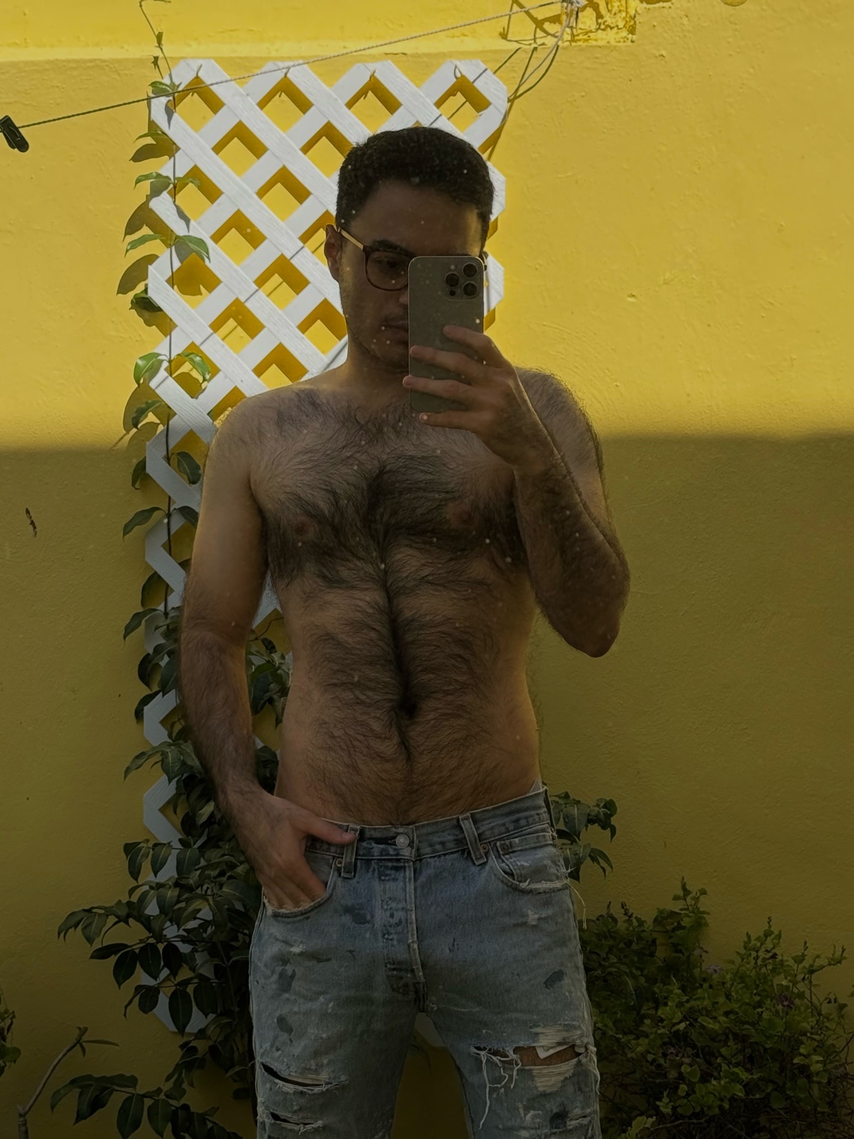 A man takes a shirtless selfie in front of a yellow wall. He is wearing jeans and glasses.