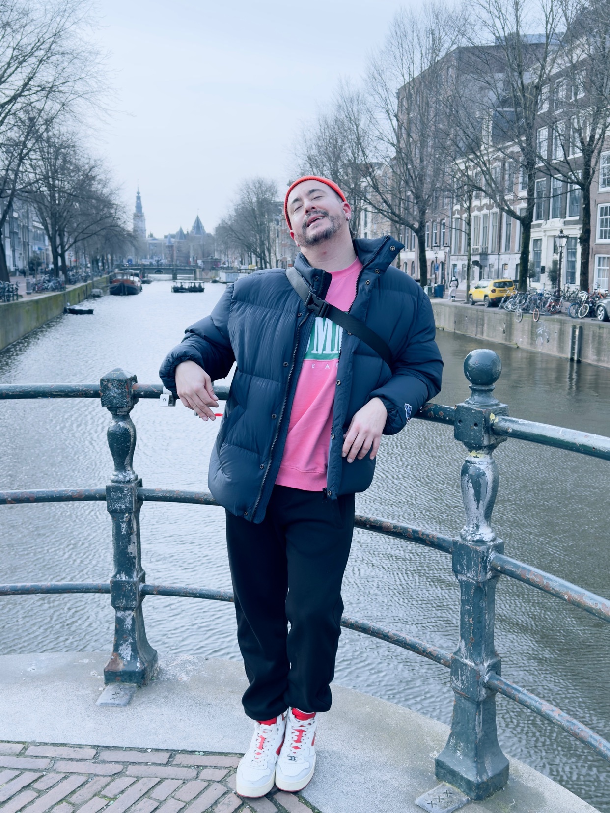 A man poses on a bridge over a canal in Amsterdam, wearing a puffer jacket and beanie.