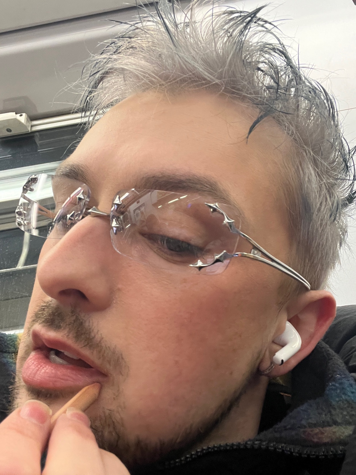 A man with silver hair and stylish glasses takes a selfie. He is wearing earpods and holding something in his mouth.