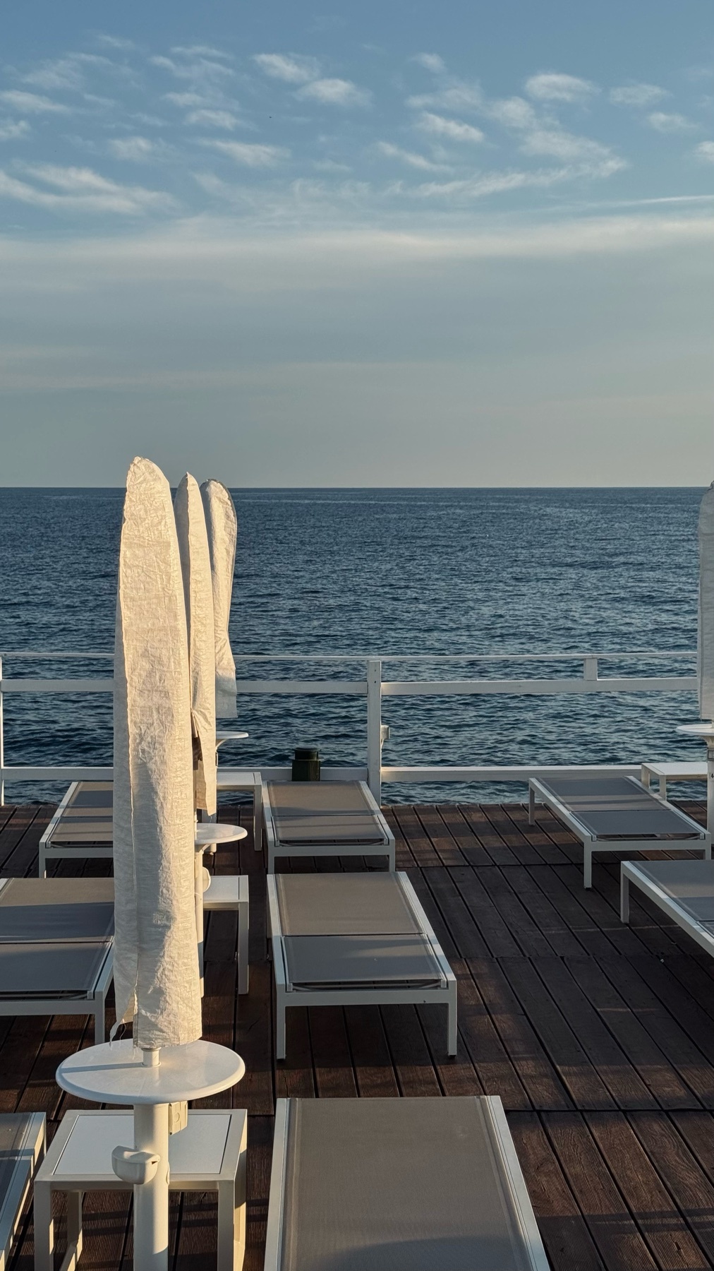 A row of empty lounge chairs and covered umbrellas sit on a wooden deck overlooking the calm blue ocean under a partly cloudy sky.