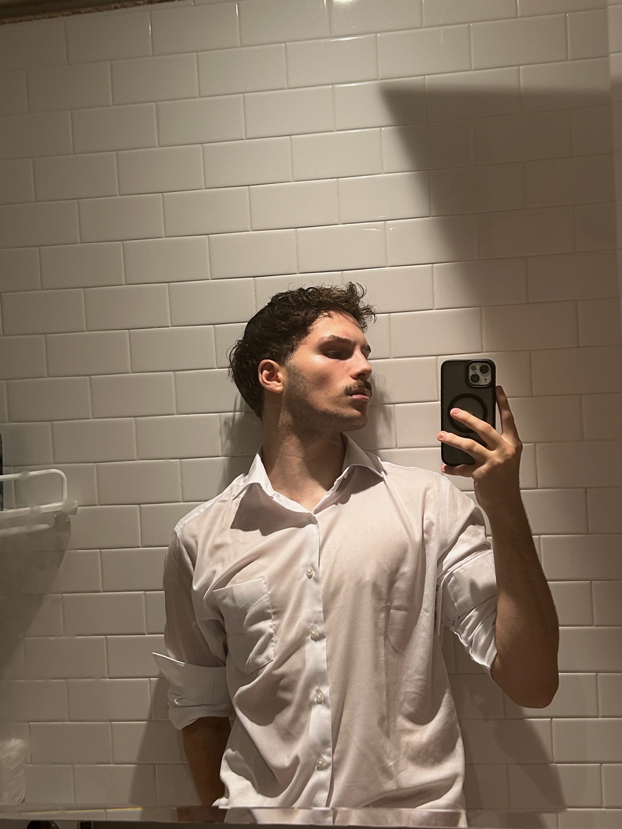 A man takes a mirror selfie in a white shirt. The photo is taken in a bathroom with white tiles.