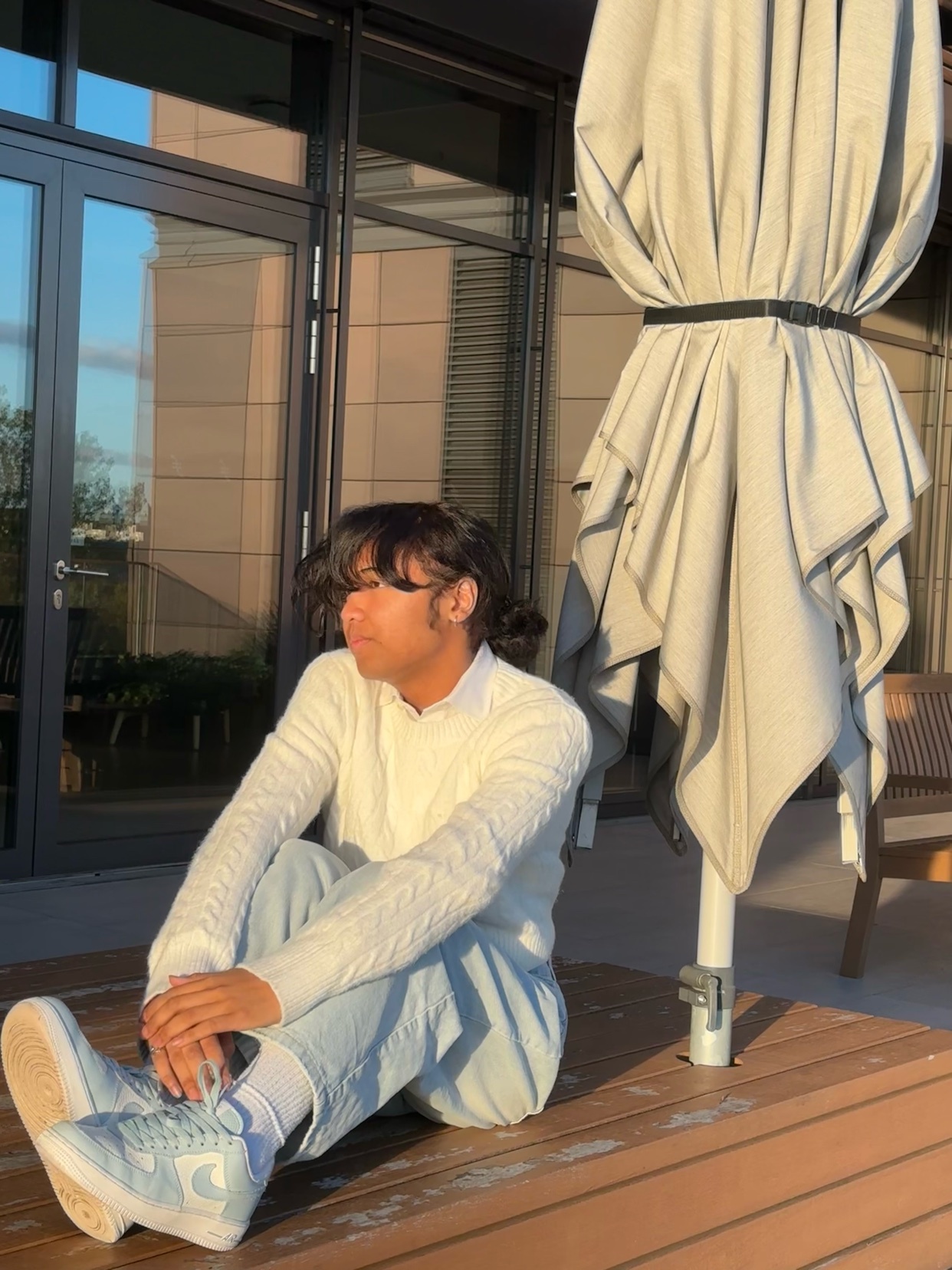A man sits on a wooden surface, wearing a white sweater and light blue jeans. The photo is taken in natural light, with a large umbrella in the background.