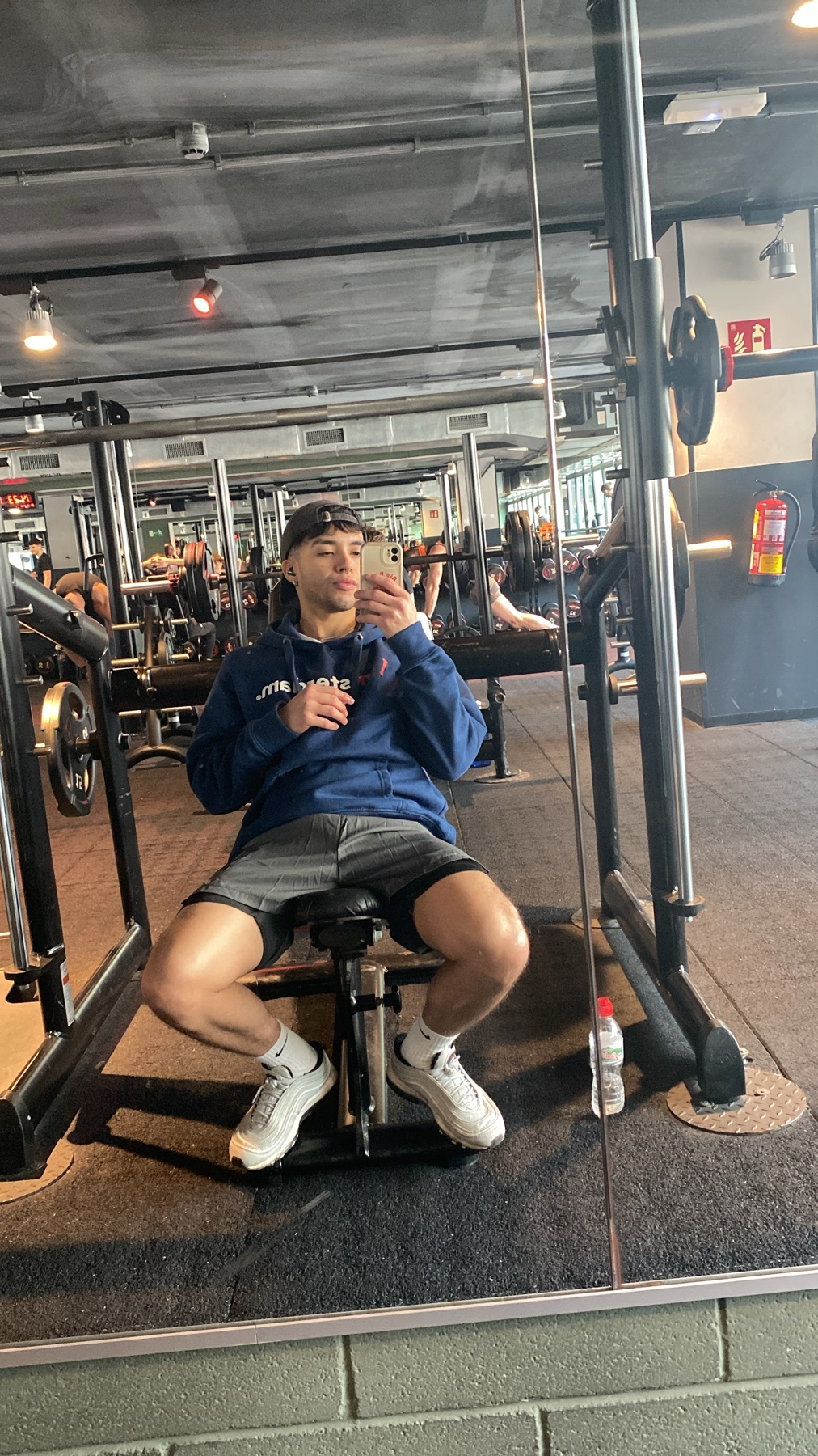 A man takes a mirror selfie at the gym. He is wearing a hoodie, shorts, and sneakers.