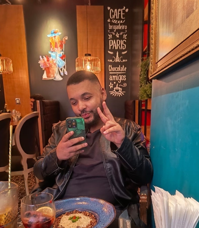 A man sits at a table in a cafe, holding his phone and making a peace sign with his fingers. A plate of food and a drink are on the table in front of him.