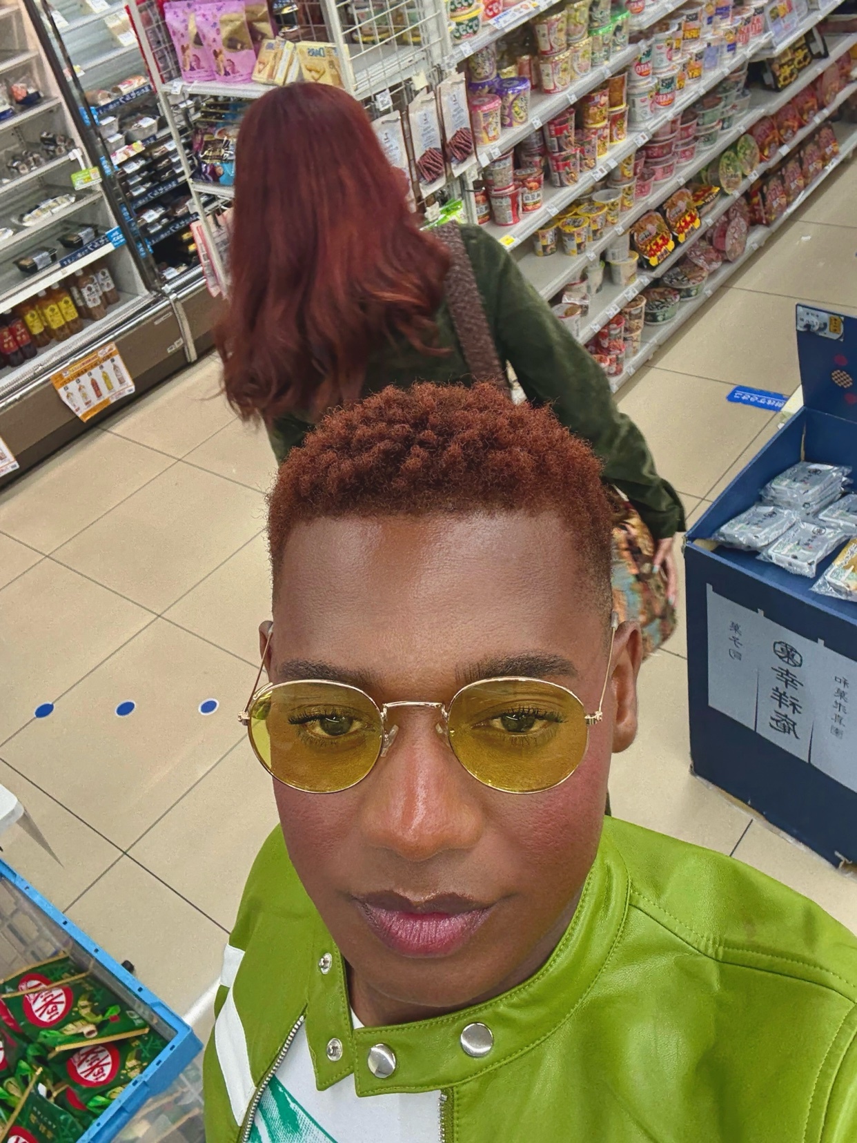 A selfie of a man wearing yellow glasses and a green jacket, with a woman with red hair in the background. They appear to be shopping in a store.
