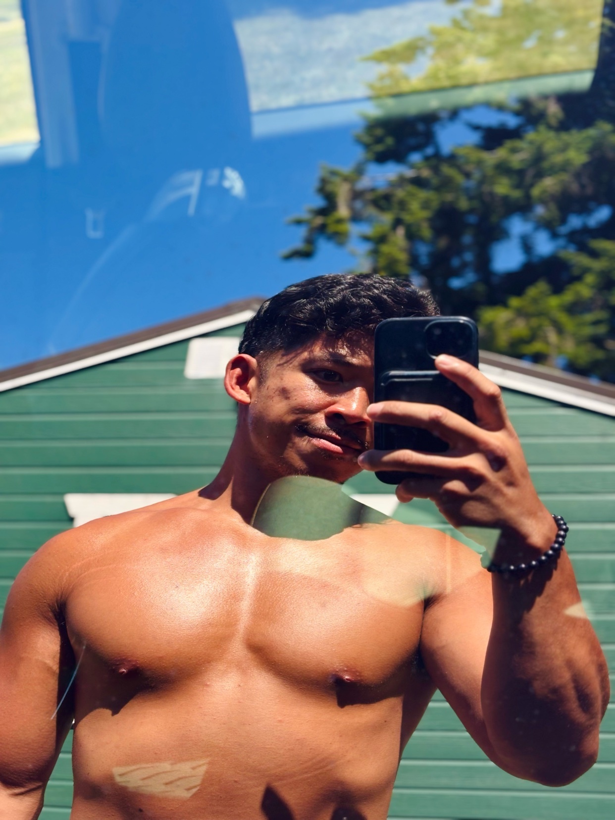 A man takes a mirror selfie, showcasing his physique. He is shirtless, flexing his arm and bicep.