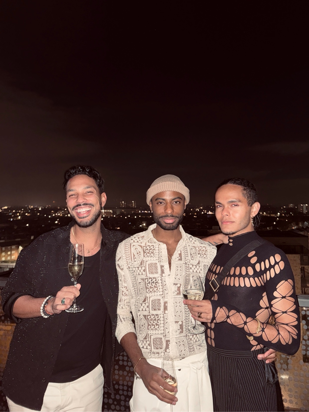 Three stylish men pose for a photo at night, holding glasses of champagne. The man on the left wears a black patterned shirt, the man in the middle wears a cream-colored patterned shirt and a beanie, and the man on the right wears a black cutout top.