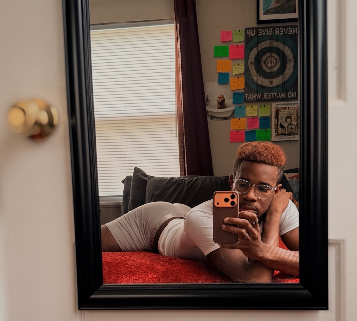 A man with red hair and glasses takes a mirror selfie while lying on a bed. He is wearing a white t-shirt and underwear.