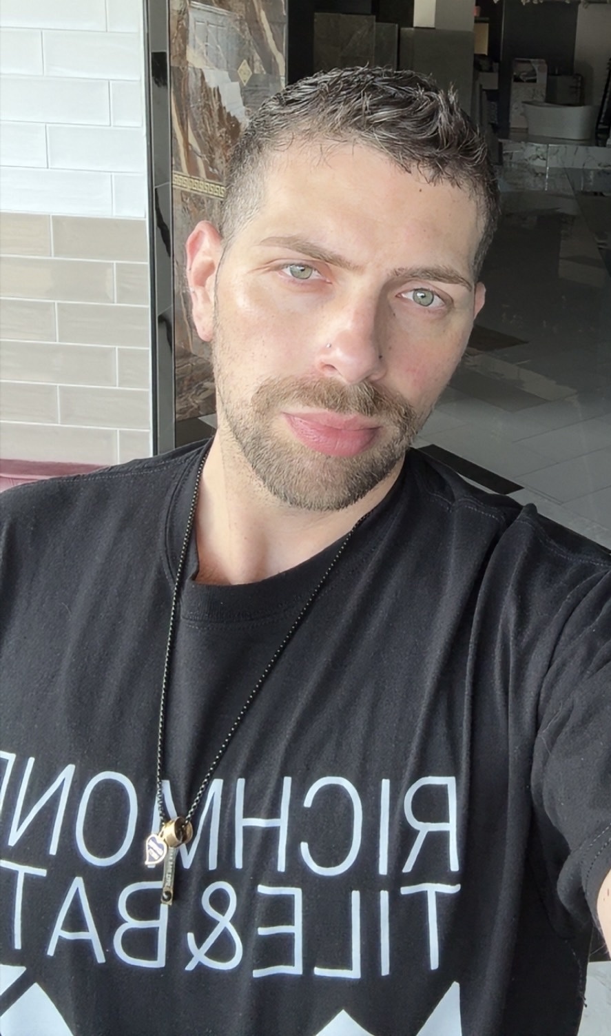 A man with short dark hair, stubble, and green eyes poses for a selfie. He is wearing a black t-shirt with white lettering and a necklace.