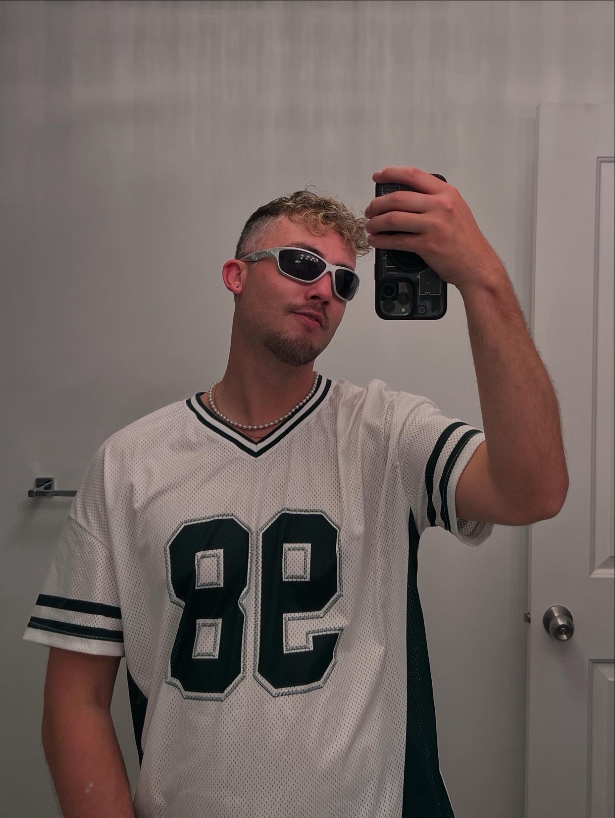 A man takes a mirror selfie. He is wearing a jersey and sunglasses.
