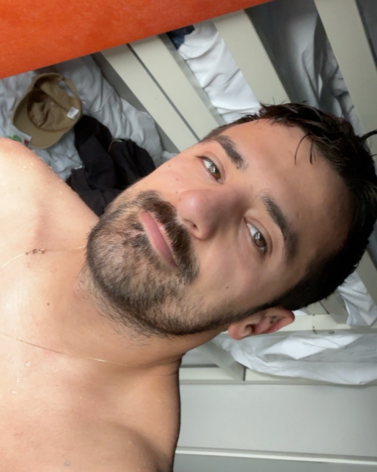 A man takes a selfie, shirtless. He has dark hair and a beard.